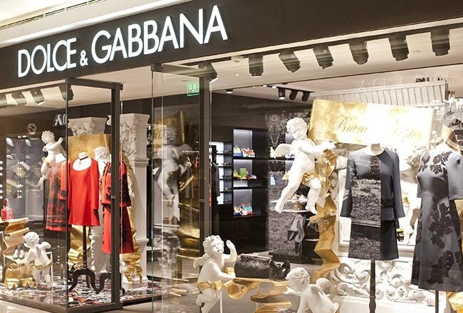 A Dolce & Gabbana store display featuring mannequins dressed in elegant black and red outfits, with decorative cherub statues and a fashionable, luxurious atmosphere.