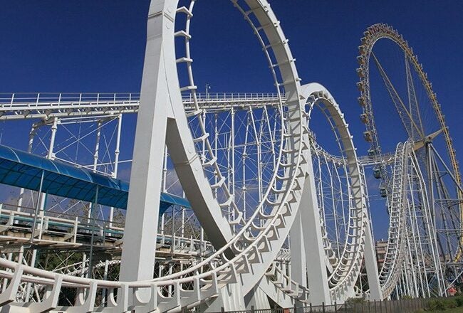 Roller coaster with multiple loops and a steep drop against a clear blue sky, surrounded by trees and a few people in the background.