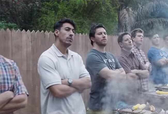 A group of people stand with their arms crossed in front of a row of barbecues, with smoke rising, suggesting a gathering or cookout.