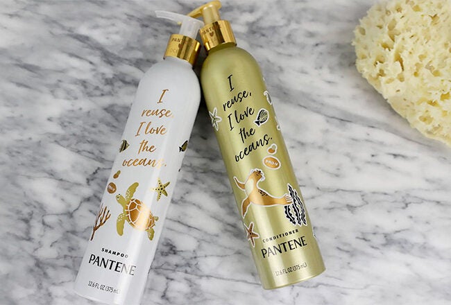 Two Pantene bottles with ocean-themed designs and the phrase "I reuse, I love the ocean" on a marble surface, next to a natural sponge.