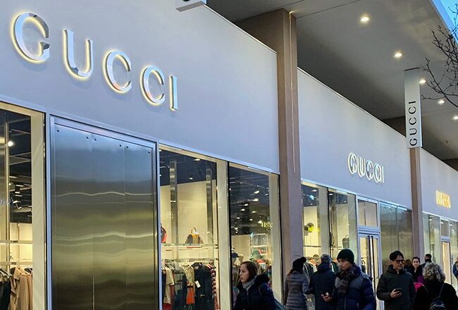 A shopping area featuring a Gucci storefront with large glass windows displaying clothing and accessories. People are walking along the sidewalk.