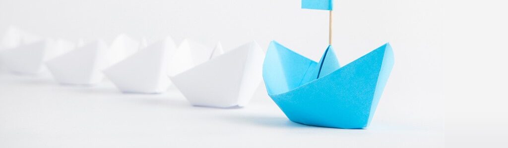 A blue origami boat with a small matching flag leads a line of white origami boats, symbolizing leadership or standing out.