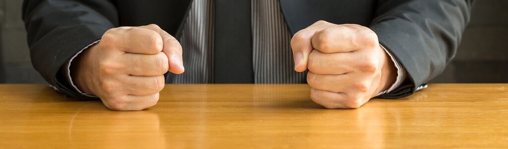 A person wearing a suit sitting at a table with both fists clenched on the surface, suggesting frustration or determination.