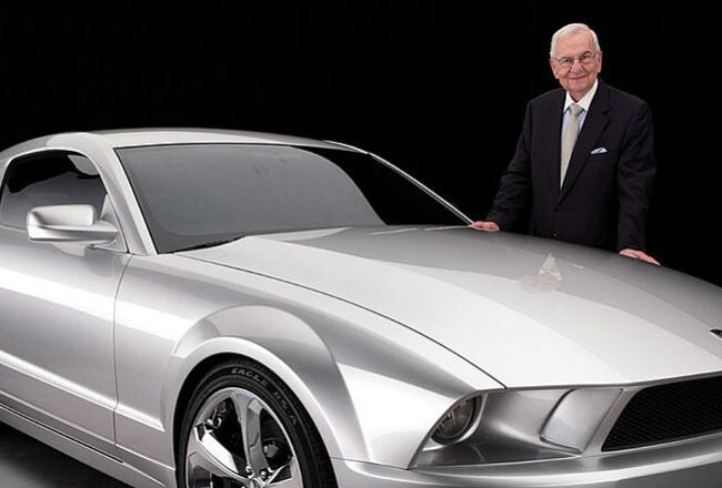 A person in a suit stands beside a sleek, silver sports car with a modern design. The background is dark, highlighting the vehicle''s glossy finish.