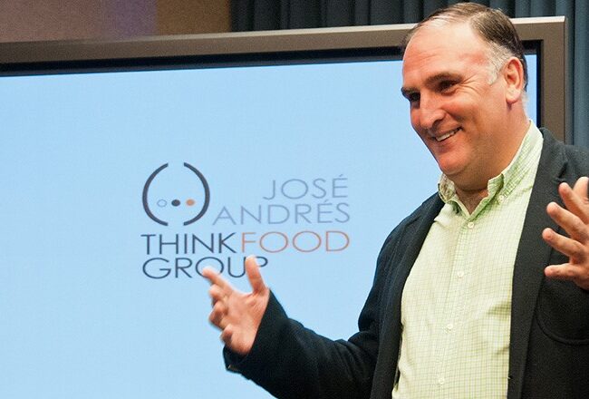 A person gesturing while standing in front of a screen displaying the logo and text: "José Andrés ThinkFood Group." An American flag is visible in the background.