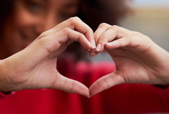 Person forming a heart shape with their hands, wearing a red sweater. The focus is on the hands, symbolizing love or affection.