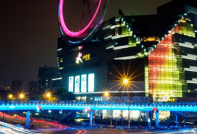 A brightly lit modern building at night, featuring a large illuminated pink circle on the roof. A light trail from moving vehicles is visible in the foreground.