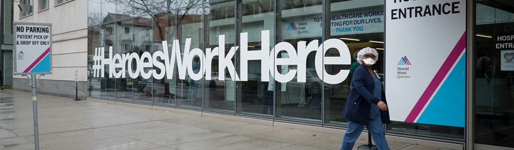 A person wearing a face mask walks past a hospital entrance with large text on the window reading "#HeroesWorkHere" and other signs nearby.