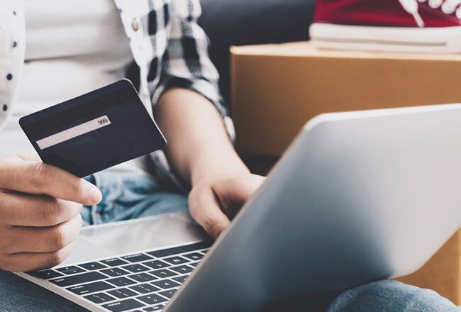 Person holding credit card while using a laptop, surrounded by delivery boxes. Represents online shopping or e-commerce.