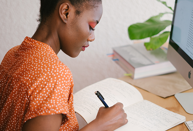 A person in a polka dot shirt writes in a notebook at a desk with a computer. The setting includes books and a plant, suggesting a study or work environment.