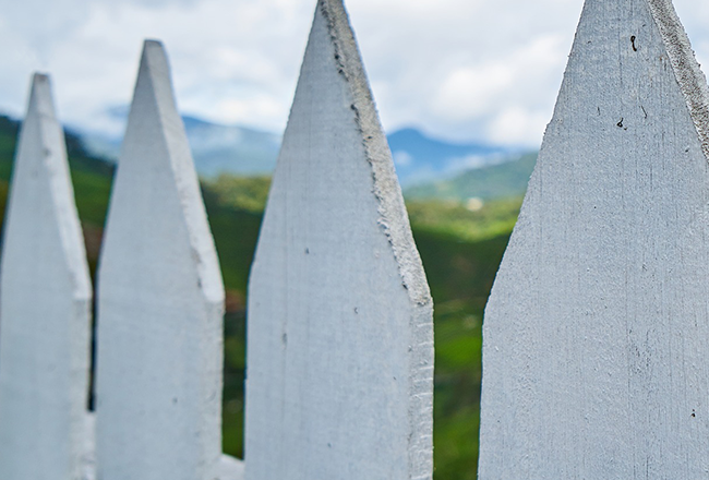 Close-up of a white picket fence with pointed tops, set against a blurred backdrop of green hills and a cloudy sky.