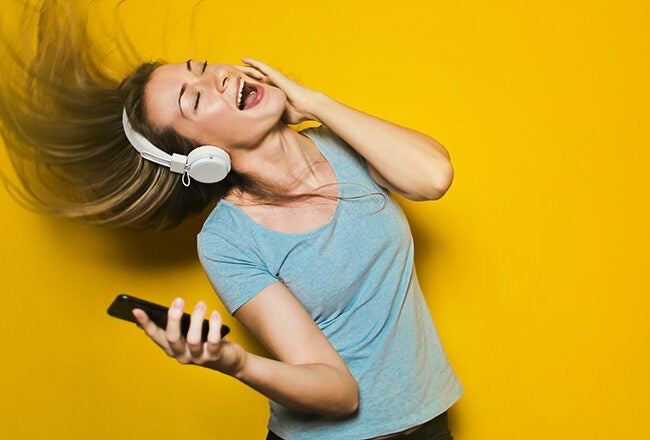 Person wearing headphones, dancing with eyes closed and hair flying, holding a smartphone in a joyful expression against a yellow background.