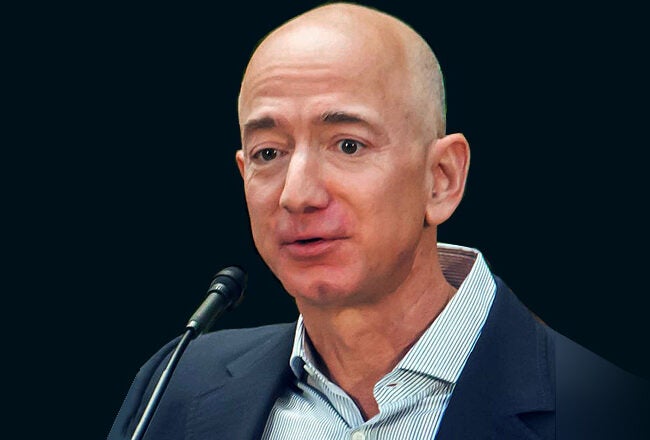A headshot of a person speaking at a microphone, dressed in a suit and collared shirt, with a neutral background.