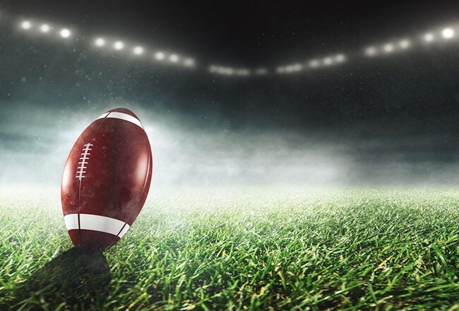 A football standing upright on a grassy field under bright stadium lights, with mist or smoke in the background.