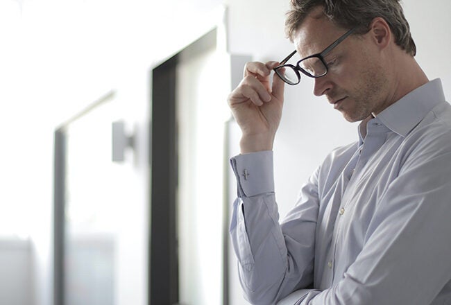 A person wearing glasses appears thoughtful or stressed, leaning against a wall in a brightly lit hallway.