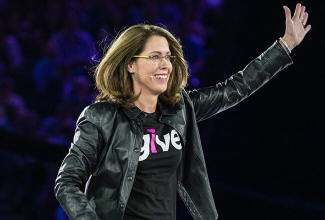 A person wearing glasses and a black leather jacket with a "give" shirt, smiling and waving on stage, addressing an audience.