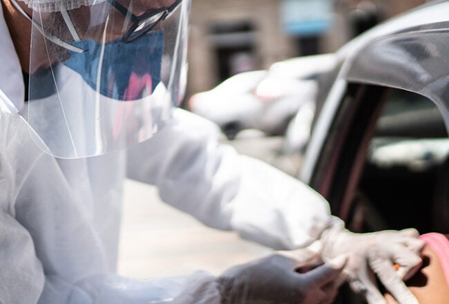 A healthcare worker wearing protective gear and a face shield administers a vaccine to a person sitting in a car during a drive-through vaccination.