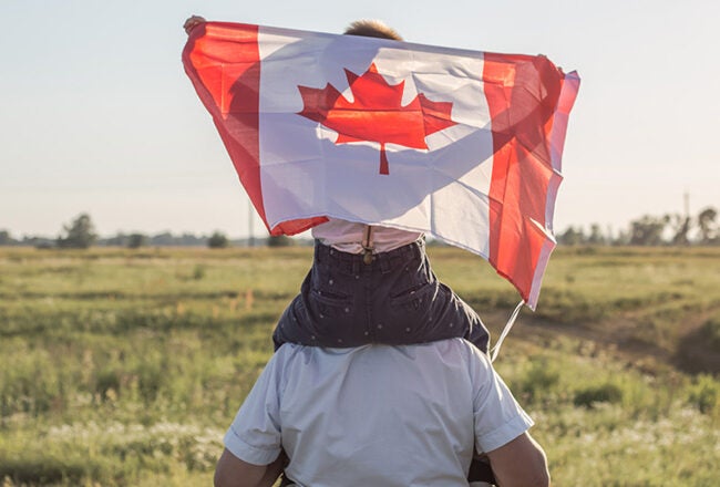 A person carrying a child on their shoulders in a field. The child is holding the Canadian flag, which is prominently displayed.