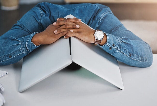 A person in a denim shirt, sitting at a desk, rests their head on a closed laptop with hands clasped on top, appearing frustrated or tired.