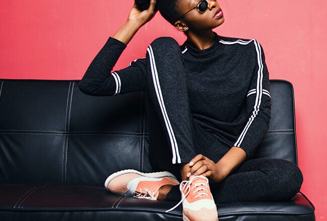 A person wearing sunglasses and a dark tracksuit sits on a black leather couch against a red wall, with one hand raised to their head.