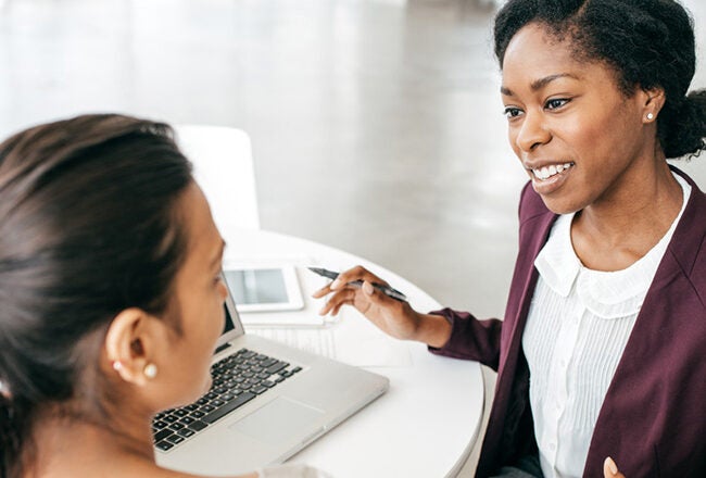 Two people sitting at a table engaged in a discussion. One is holding a pen, and a laptop is open in front of them.