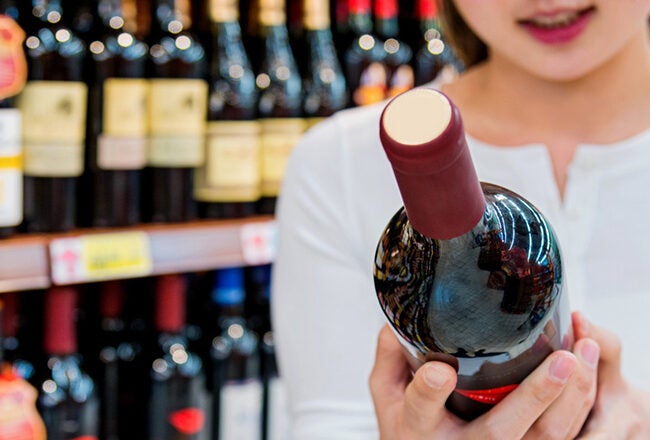 A person in a white shirt is holding a wine bottle in a store aisle with shelves lined with various wine bottles.