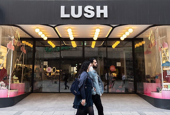 The image shows a storefront of Lush, a cosmetics retailer. Two people are walking in front of the store, which has large glass windows displaying products.