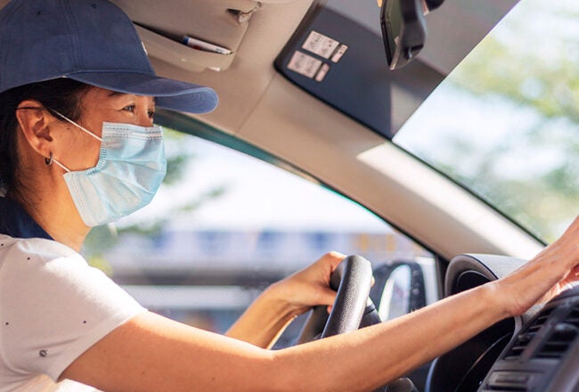 A person wearing a face mask and cap drives a car, using a GPS device mounted on the dashboard.