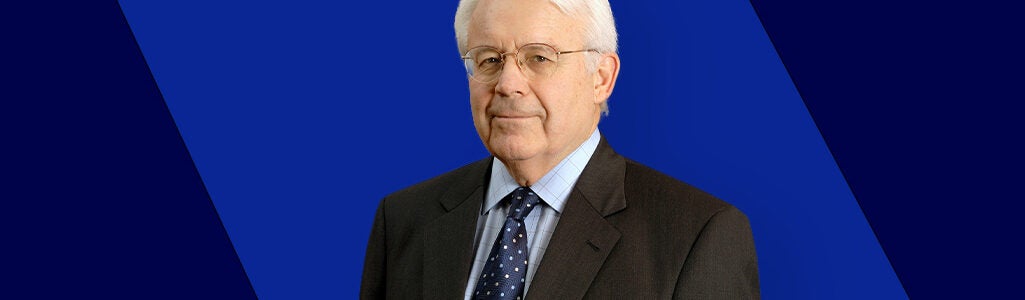 A headshot of an older individual wearing a suit and tie against a blue geometric background.