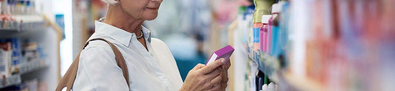 An older adult with white hair is shopping in a store aisle, examining a product held in one hand. The setting appears to be a pharmacy or health store, with shelves of various items visible. They are wearing a light blouse and have a large tan