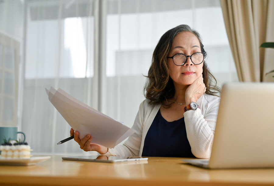 Older woman looking thoughtfully at her computer and documents thinking about retirement pension cuts and other generation gap issues