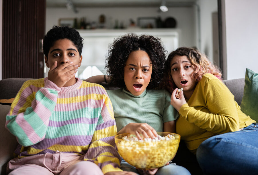 Three friends watching a movie on the couch with a bowl of popcorn and reacting with shock at what's on screen as they experience the "so bad it's good" phenomenon