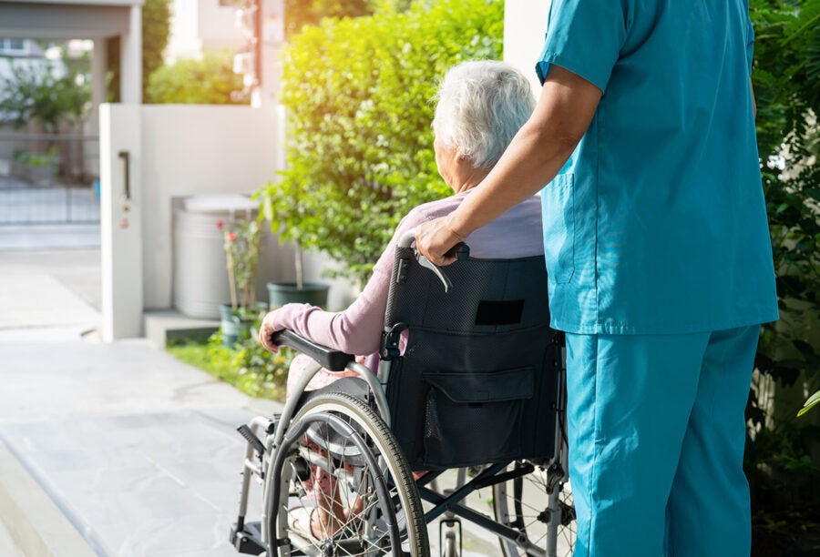 A older woman in a wheelchair being rolled along the sidewalk by a nursing home assistant