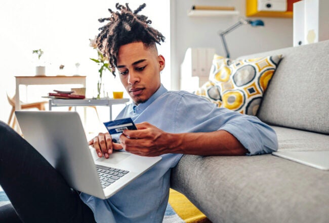 Young man looking at his credit card while on his laptop to show how AI is affecting financial well-being