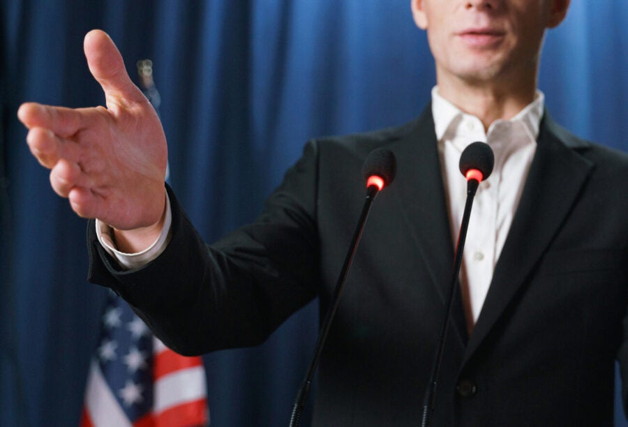 Political candidate gesturing with his hand as he speaks at a podium in front of the American flag to show how tone of voice and speech styles can influence voters