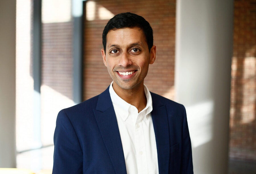 A person in a blue suit and white shirt is smiling in a headshot. The background features a brick wall and soft lighting.