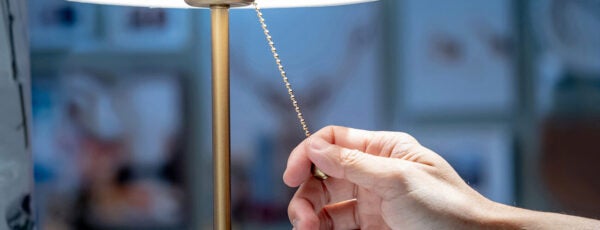 A hand pulling the chain of a lit table lamp with a pleated white shade. The background is softly blurred, suggesting an indoor setting.