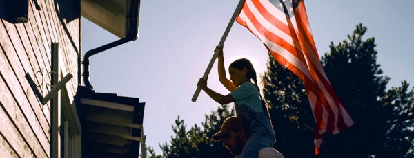 A father carrying his daughter on his shoulders as she raises an American flag to place on the outside of their house.