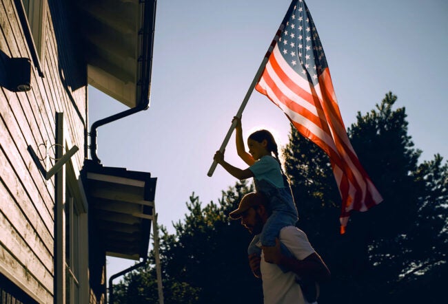 A father carrying his daughter on his shoulders as she raises an American flag to place on the outside of their house.