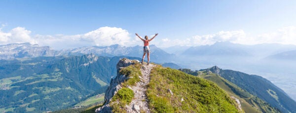 Person standing on a grassy mountaintop with their arms raised against the blue sky