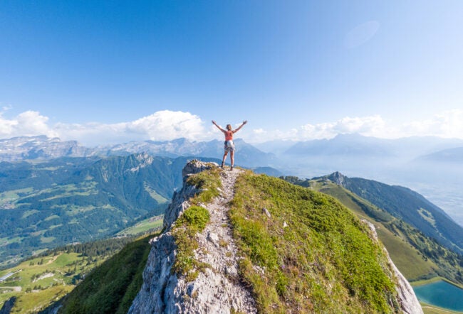 Person standing on a grassy mountaintop with their arms raised against the blue sky