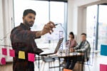 Man drawing an upward arrow on a clear board with his coworkers in the background to show why the most successful companies are scalable