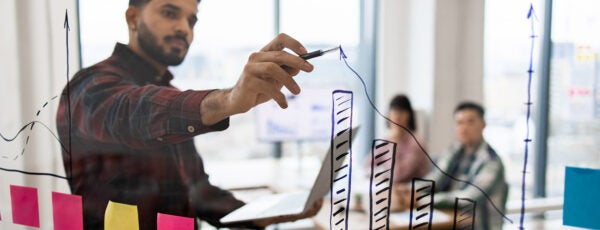 Man drawing an upward arrow on a clear board with his coworkers in the background to show why the most successful companies are scalable