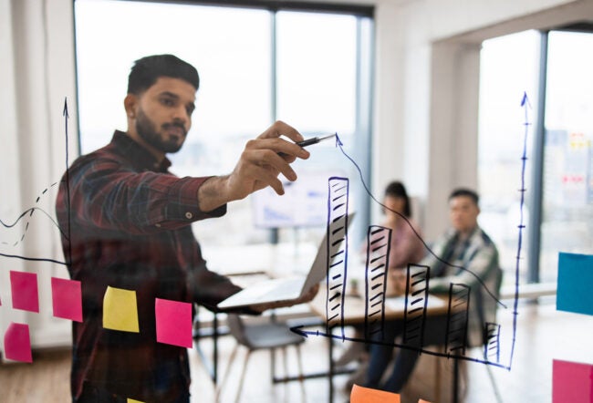 Man drawing an upward arrow on a clear board with his coworkers in the background to show why the most successful companies are scalable