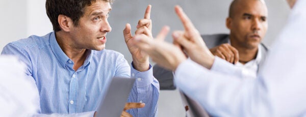 A group of people engaged in a discussion at a meeting table with laptops and documents. One person is gesturing, indicating an animated conversation.