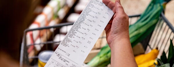 A person is holding a long grocery receipt while standing near a shopping cart filled with fresh vegetables and a pineapple.