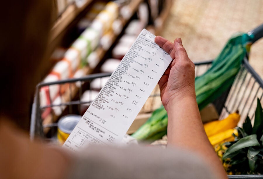 A person is holding a long grocery receipt while standing near a shopping cart filled with fresh vegetables and a pineapple.