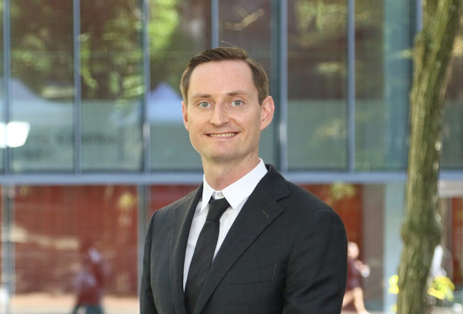 A person in a formal suit is smiling, standing outdoors with a modern glass building in the background.