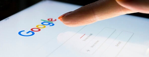 A hand tapping on a tablet with the Google search page displayed. Why does Google dominate the search engine market?