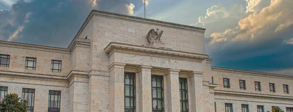 A stately government building with a classical facade and a flagpole with two flags flying. Sun rays and clouds are visible in the sky above.